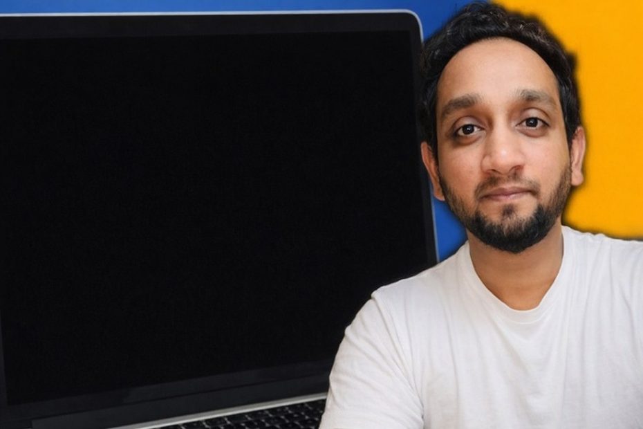 En man med skägg i vit t-shirt framför en svart laptopskärm.